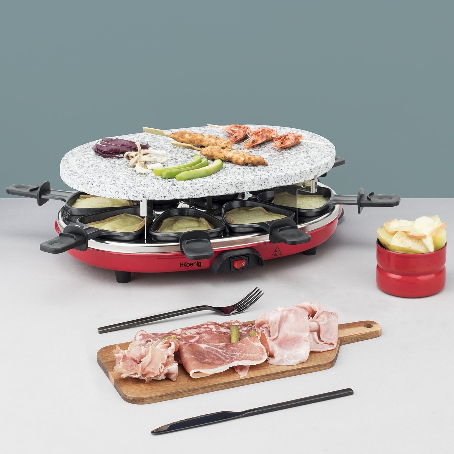 Grill per raclette con cibo: verdure, spiedini di gamberi e carne. Un tagliere con salumi e una ciotola rossa con fette di patate.