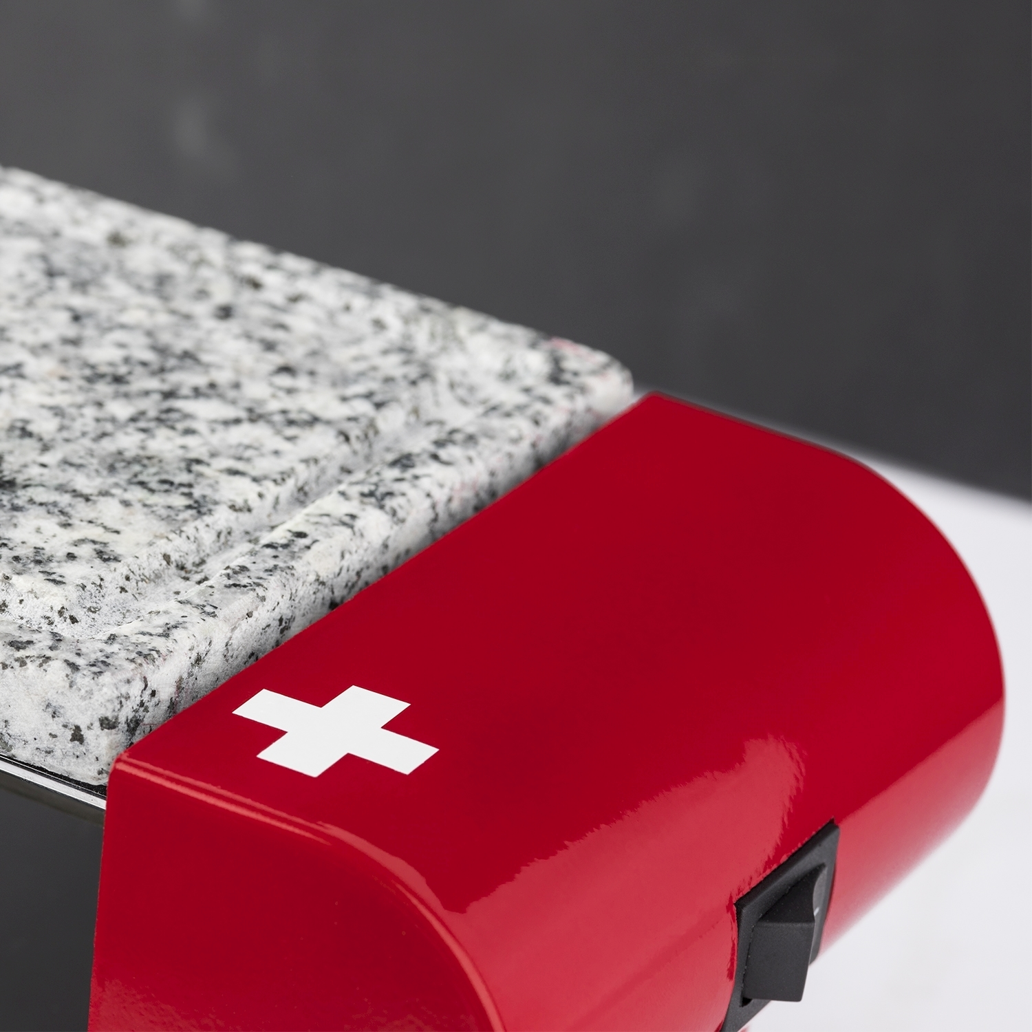 Un oggetto rosso con una croce bianca e un interruttore nero, accanto a una superficie di granito. Lo sfondo è grigio e bianco.