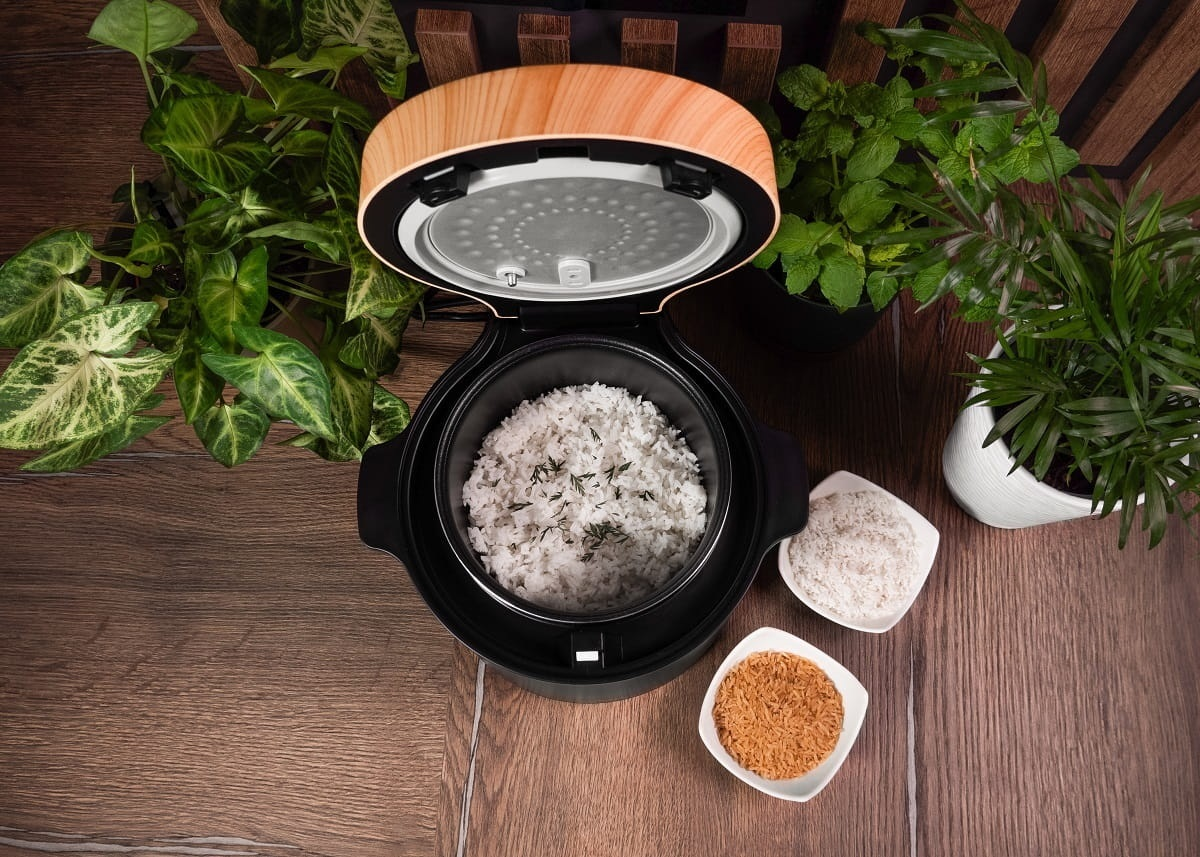 Ryżowar z ugotowanym ryżem, miskami z ryżem i roślinami na drewnianej powierzchni. Ryżowar jest otwarty, pokazując ryż.