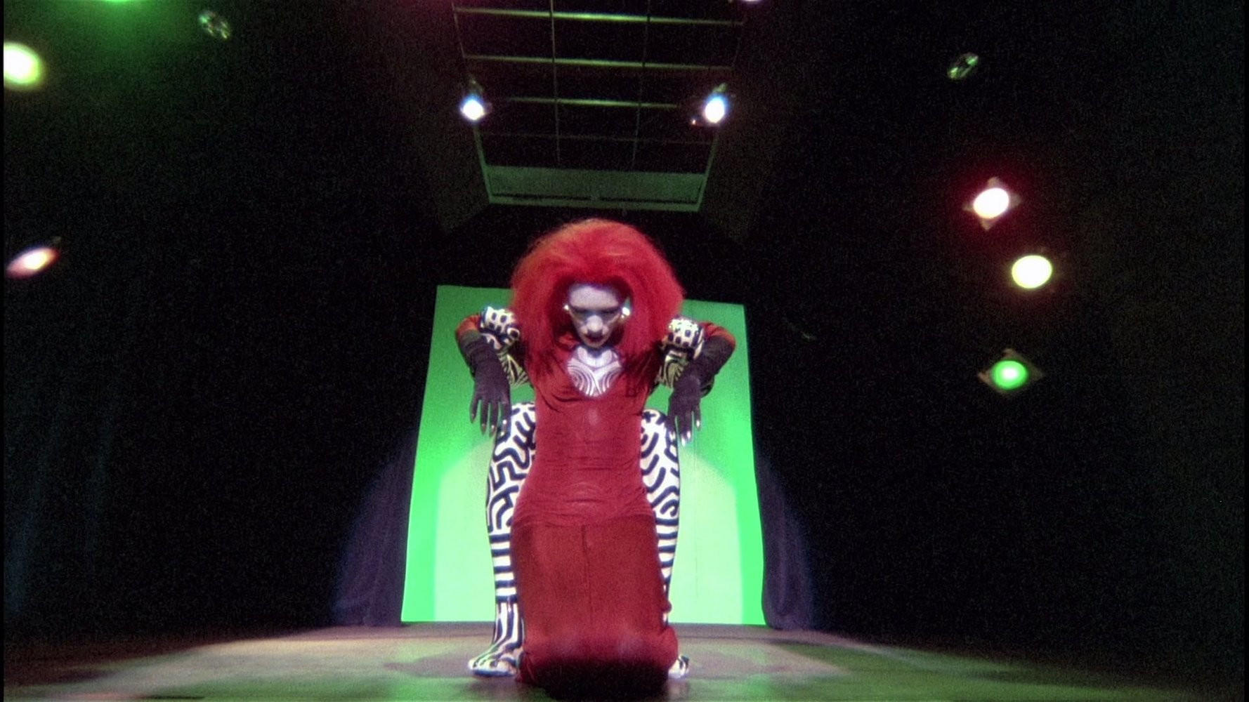 Eine Dragqueen in rotem Kleid und Perücke posiert auf der Bühne mit Hosen im Zebrastreifenmuster. Grüner Hintergrund und Bühnenbeleuchtung erhellen die Szene.