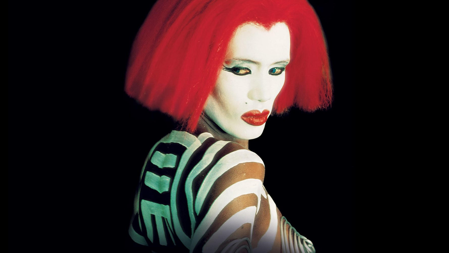 Eine Person mit rotem Haar, weißem Gesichts-Make-up und Zebrastreifen-Körperbemalung vor schwarzem Hintergrund.