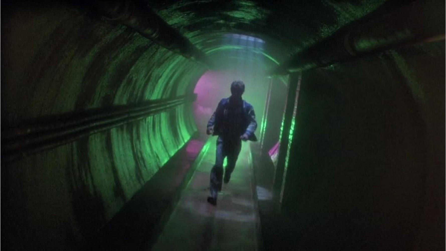 Ein Mann rennt durch einen grün beleuchteten Tunnel. Rohre verlaufen an der Decke. Lila und grüne Farbtöne erhellen die Szene.