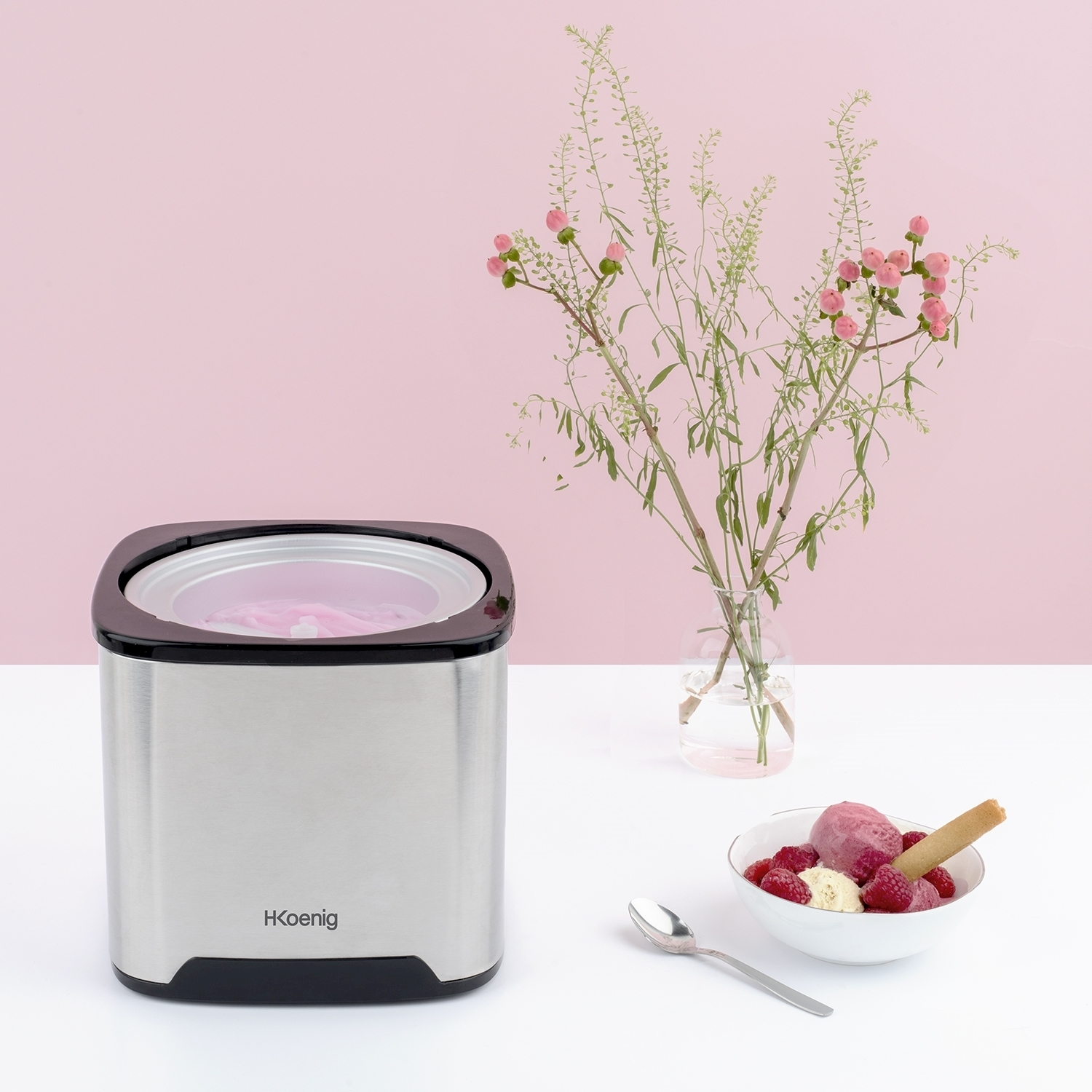 Una gelatiera, una ciotola di gelato con lamponi e un wafer, fiori in un vaso, su un tavolo bianco, sfondo rosa.