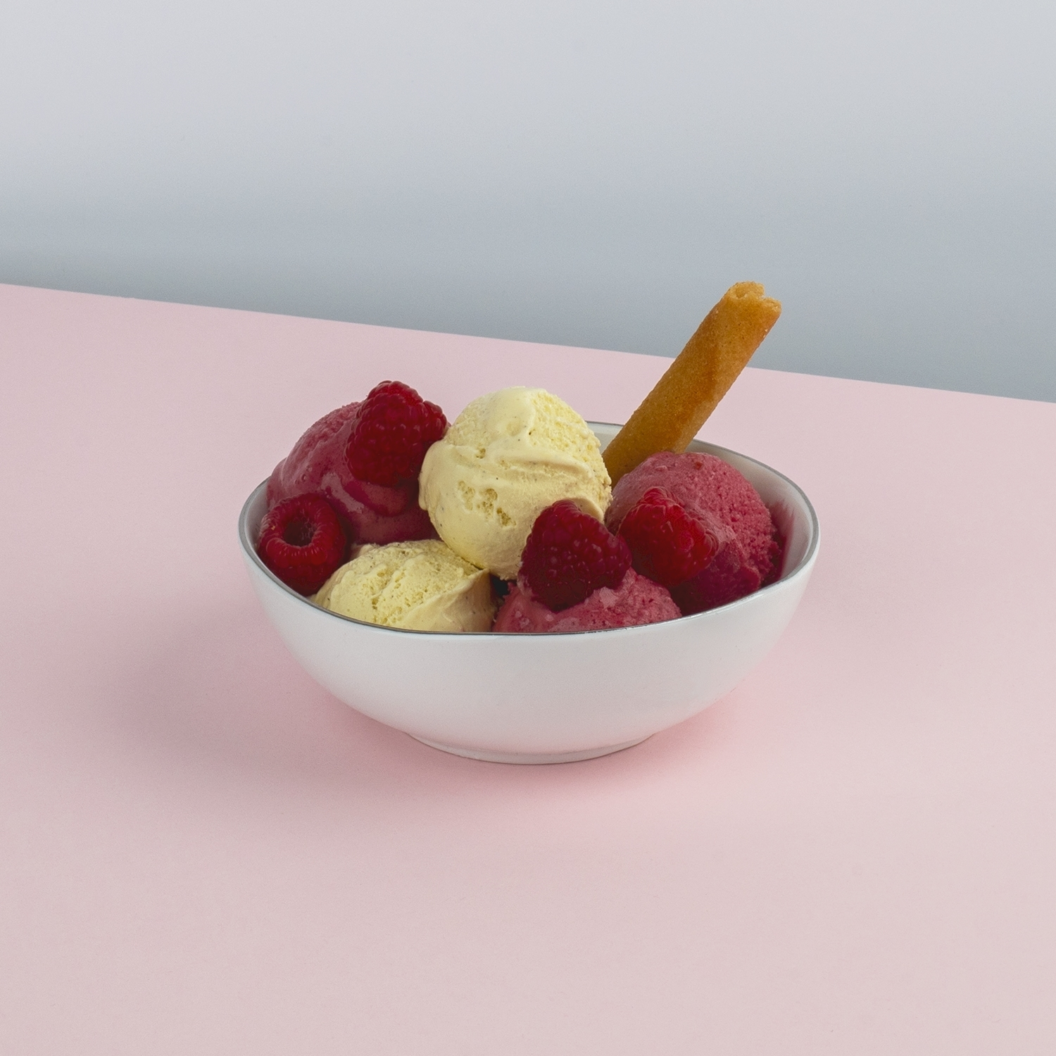 Una ciotola bianca contiene palline di gelato con lamponi e un wafer. Lo sfondo è rosa e grigio chiaro.