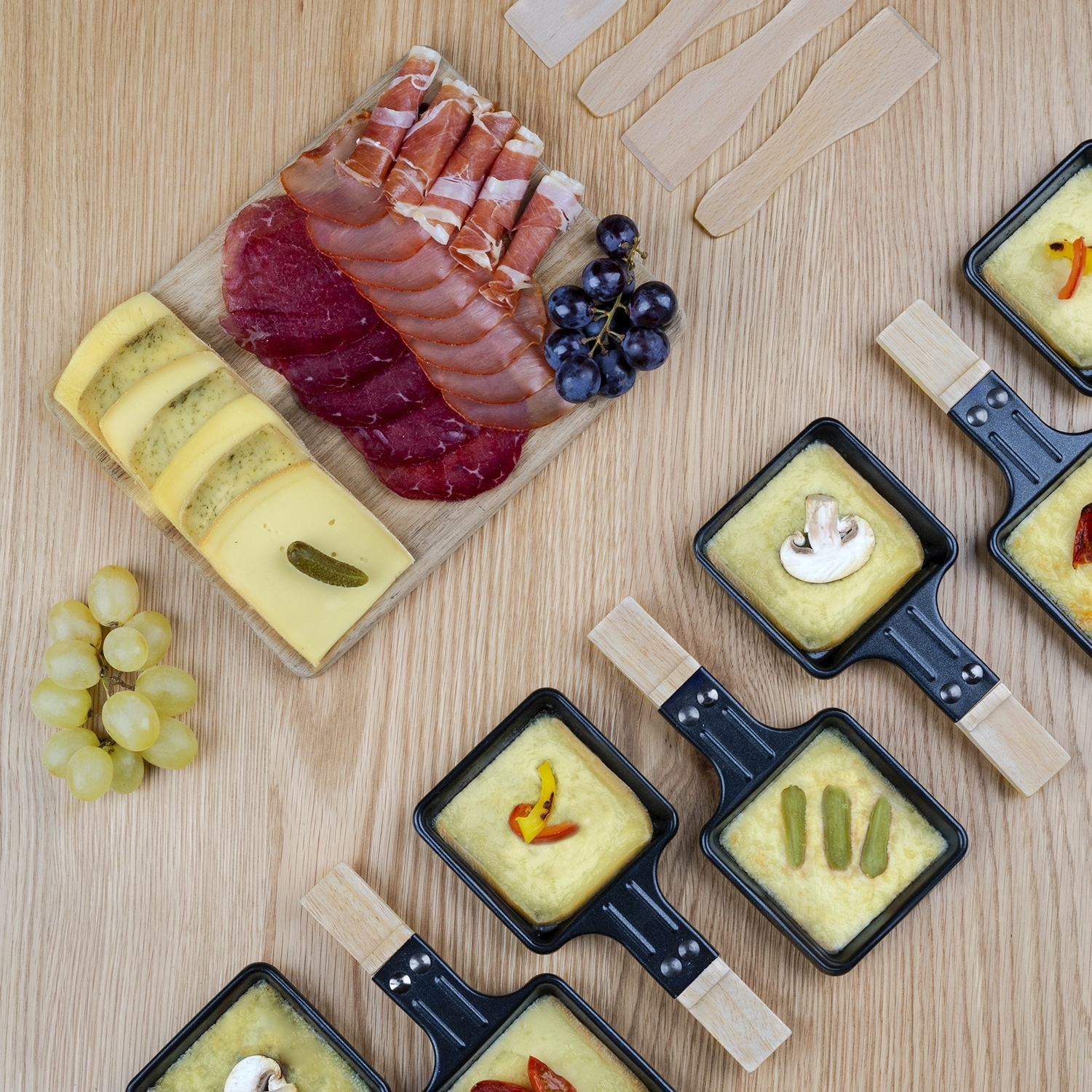 Tavolo in legno con formaggio, salumi, uva e piccole padelle di formaggio fuso con condimenti. Sono presenti anche spatole di legno.