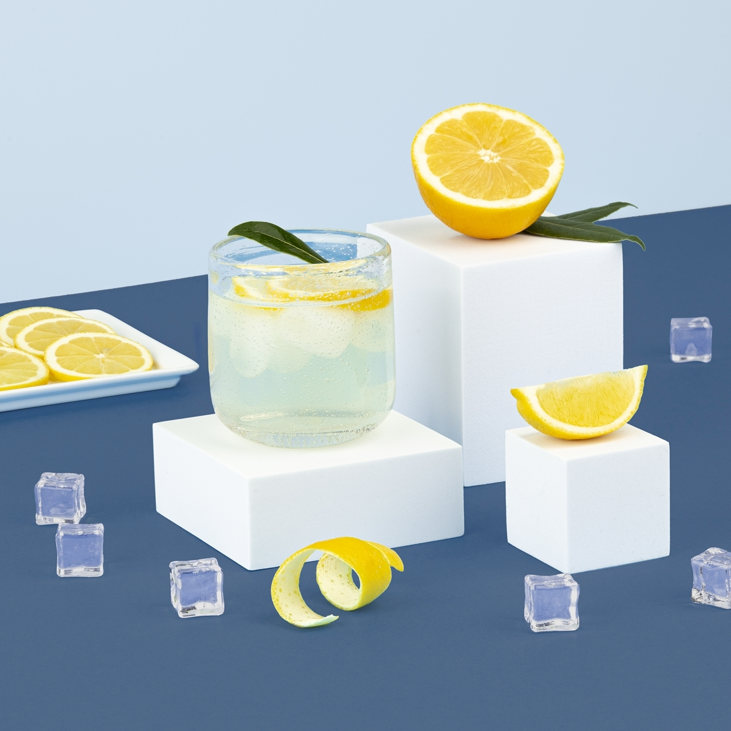 Una bevanda rinfrescante al limone con cubetti di ghiaccio e fette di limone su blocchi bianchi su sfondo blu.