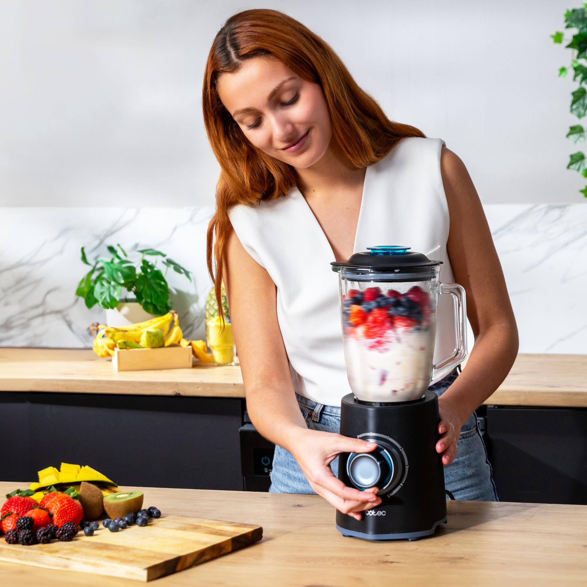 Mujer haciendo un batido con una licuadora. Frutas como fresas, arándanos y kiwi están en la mesa.