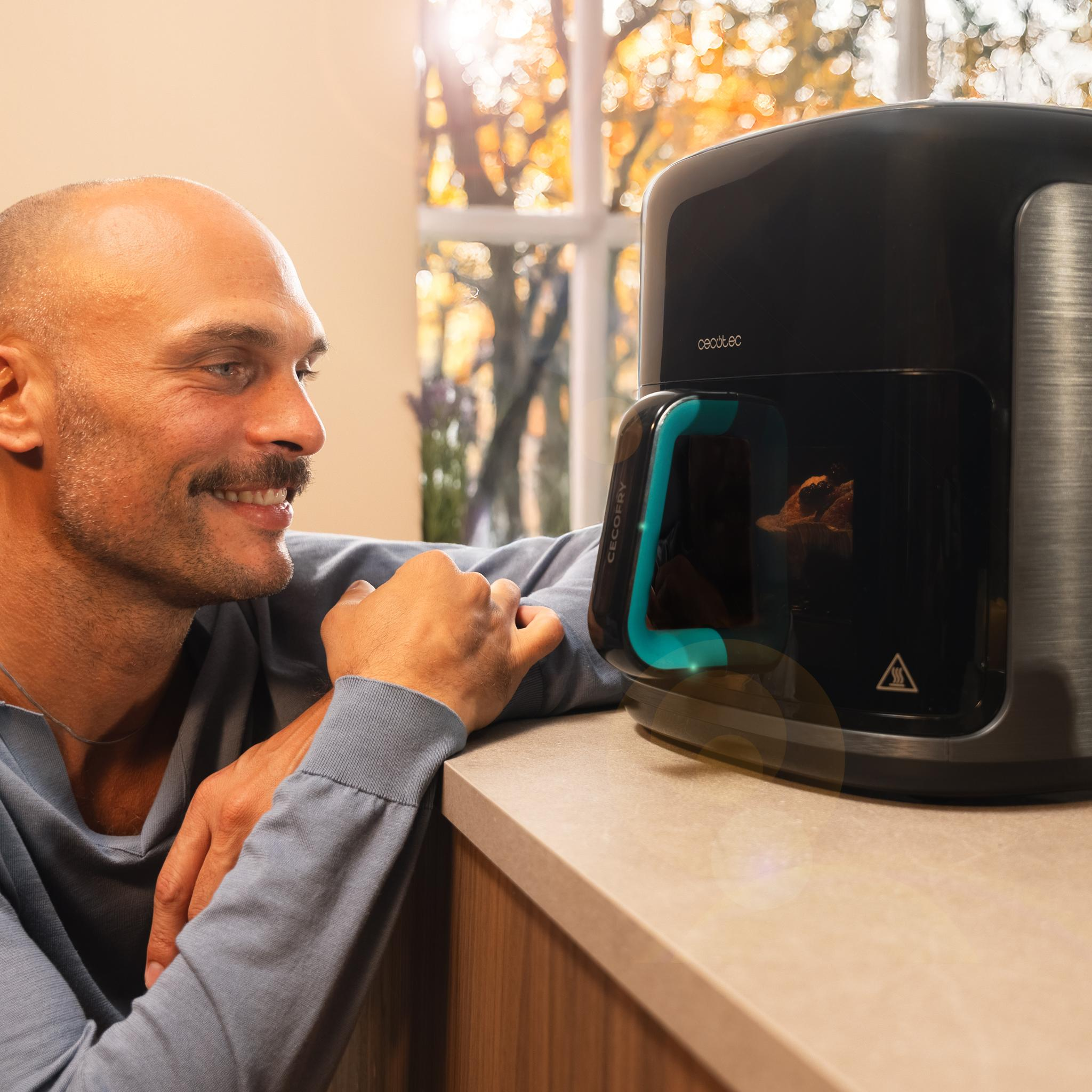 Un hombre sonríe, mirando una cafetera negra Cecotec con un asa verde azulado. El fondo muestra una ventana con follaje otoñal.