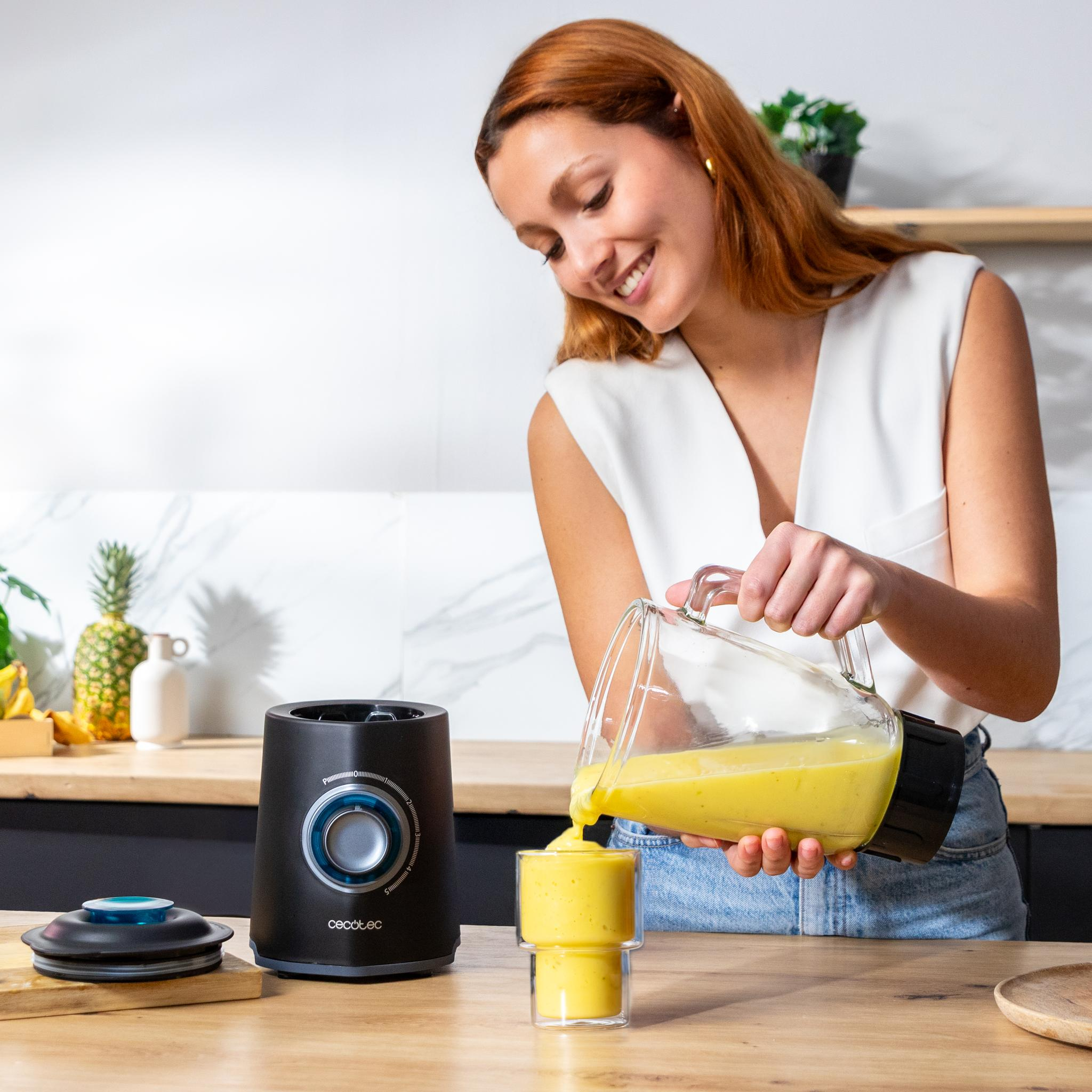 Mujer vertiendo batido amarillo en un vaso. Una licuadora está en el mostrador. Piña y otras frutas al fondo.