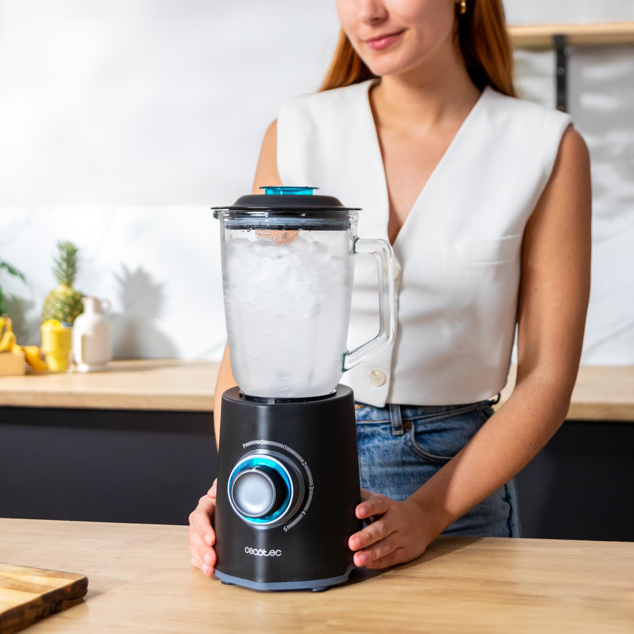 Mujer con una batidora. La batidora es negra y tiene un recipiente de vidrio lleno de agua. Ella está sonriendo.