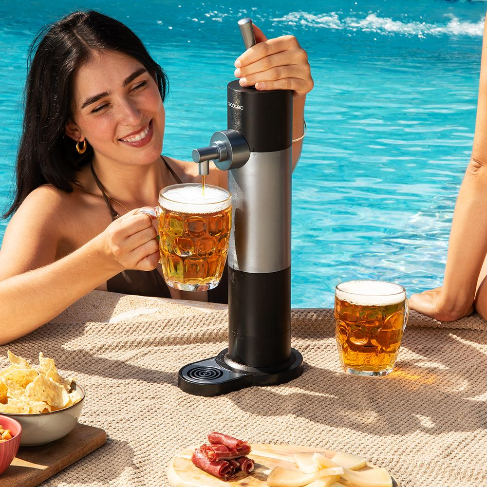 Mujer en piscina sirviendo cerveza. Hay aperitivos y otra jarra de cerveza en la mesa. El fondo es una piscina.
