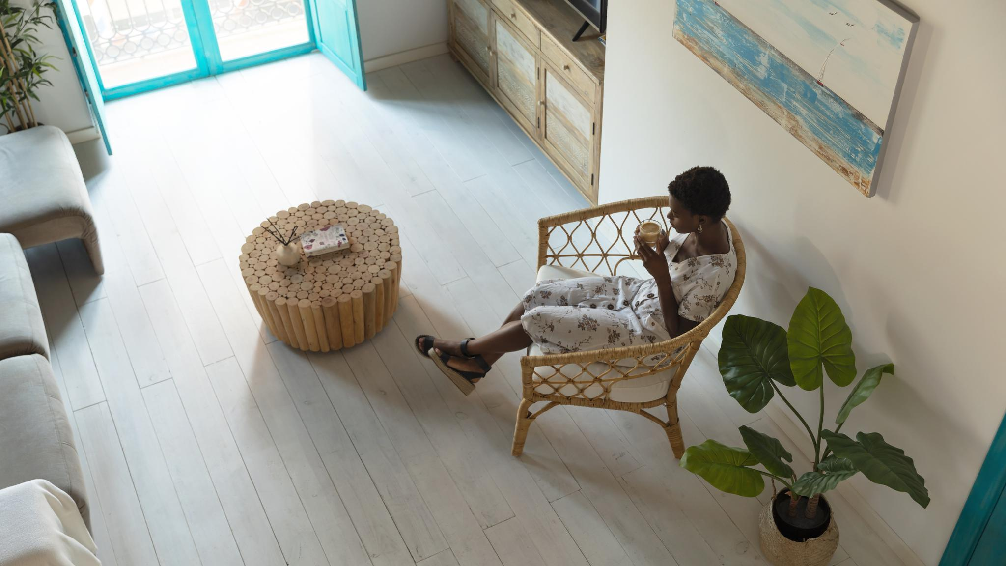 Mujer con vestido floral sentada en silla de mimbre, bebiendo café. Mesa de madera y planta grande al frente. Habitación luminosa.