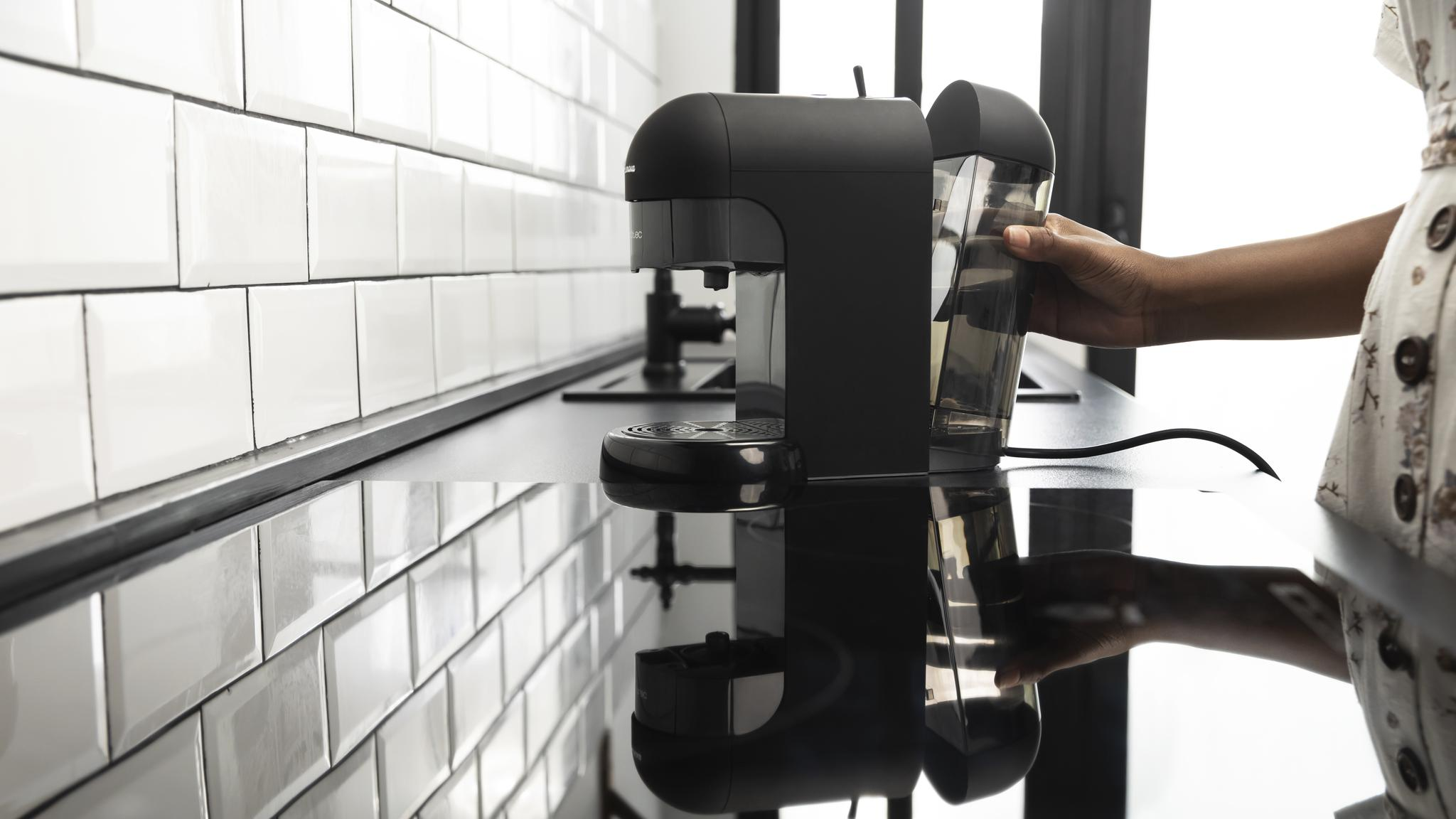 Una cafetera negra sobre una superficie negra. Una persona sostiene un recipiente de agua. Pared de azulejos blancos en el fondo.