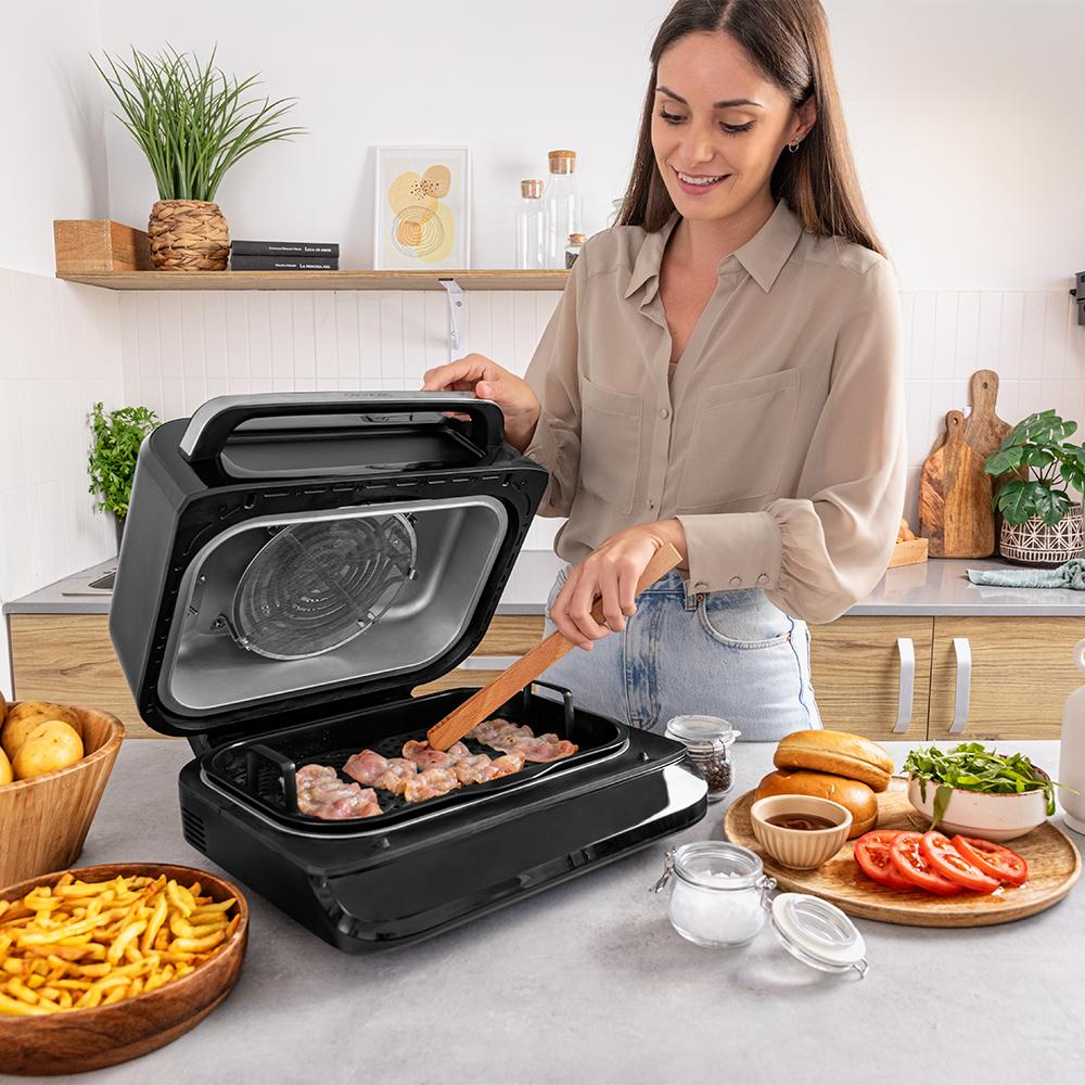 Mujer cocinando en una cocina. Usa una parrilla. Hay papas fritas, tomates, panecillos y ensalada en la encimera.