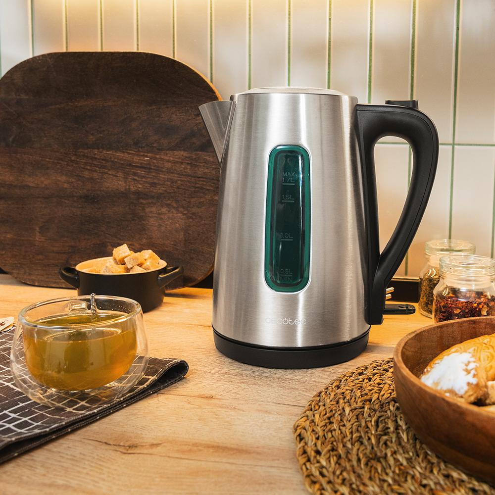 Una tetera eléctrica de acero inoxidable con asa negra, un vaso de té y alimentos sobre una mesa de madera.