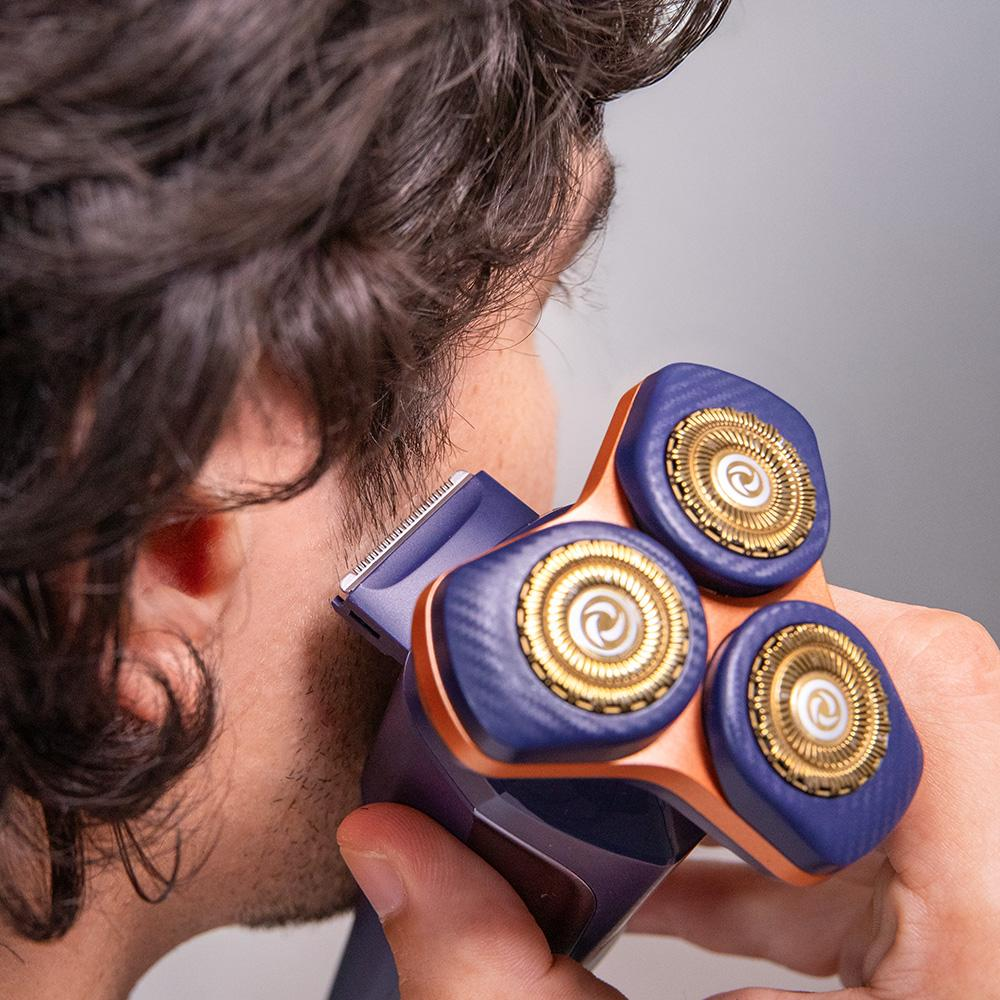 Un hombre se afeita la barba con una afeitadora eléctrica. La afeitadora es azul, naranja y dorada. El fondo es gris.