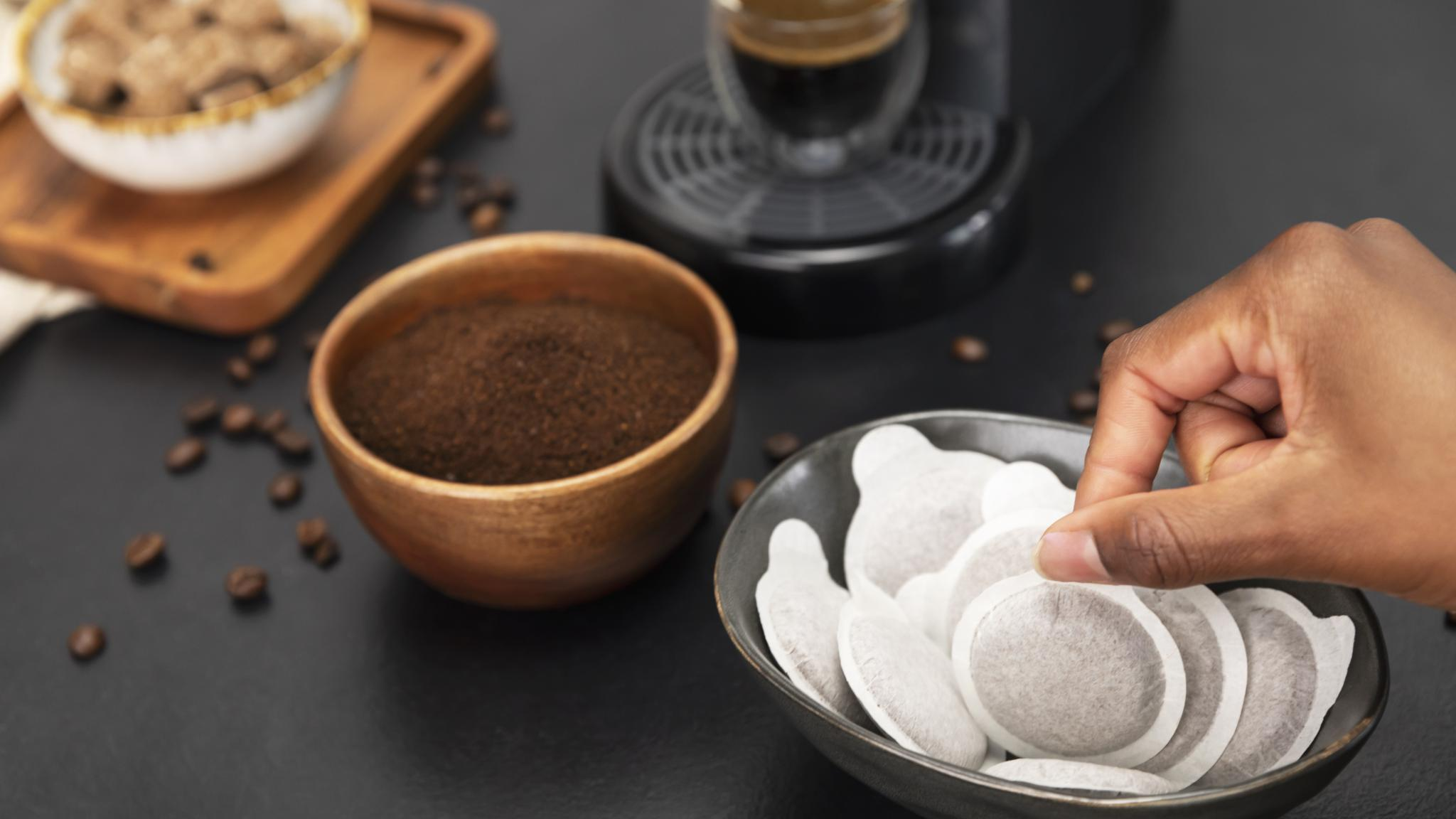 Una mano alcanza las cápsulas de café en un cuenco. También hay cuencos con café molido, granos y una cafetera.