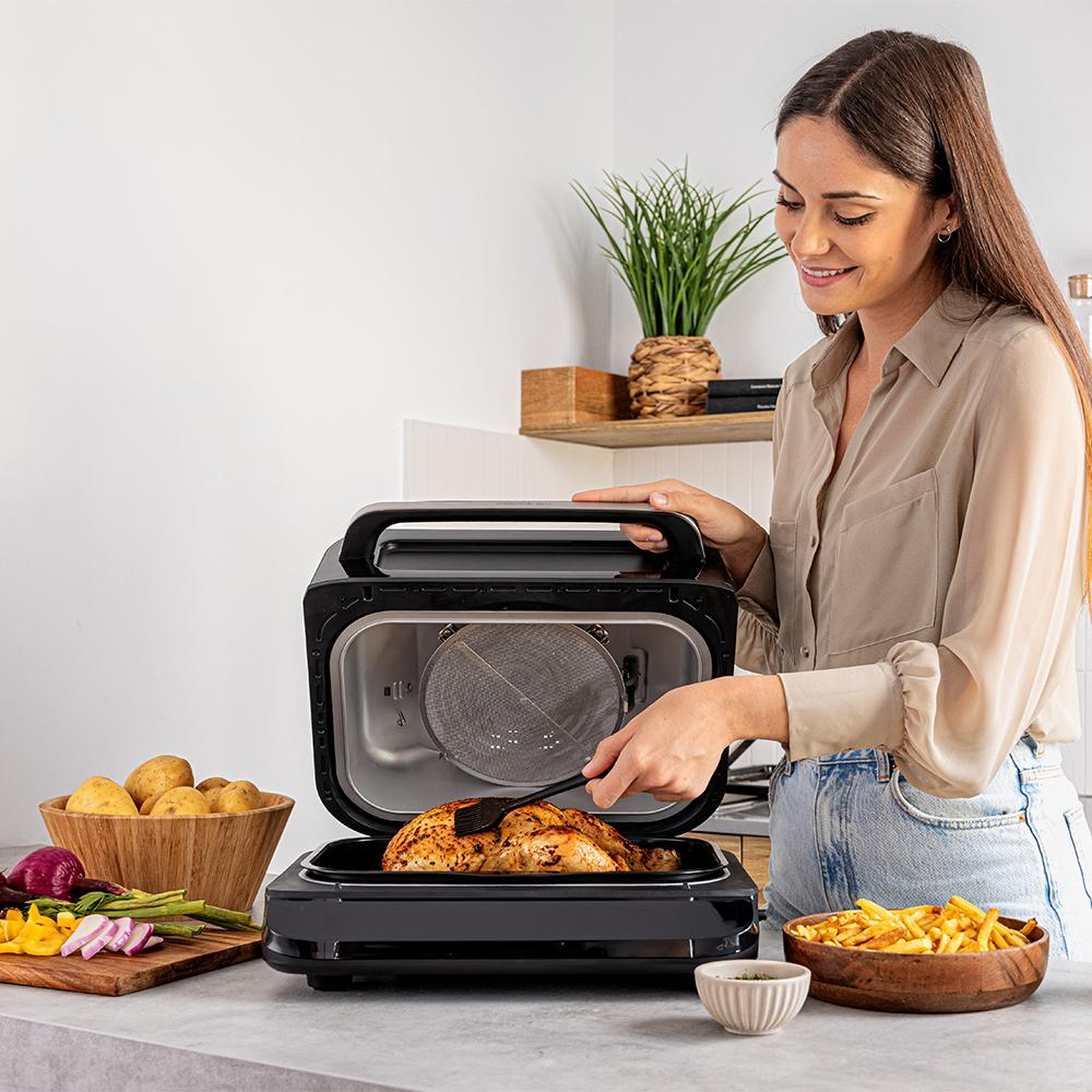 Mujer usa freidora de aire. Pollo dentro. Tazón de papas fritas a la derecha. Papas y verduras a la izquierda. Planta en el fondo.