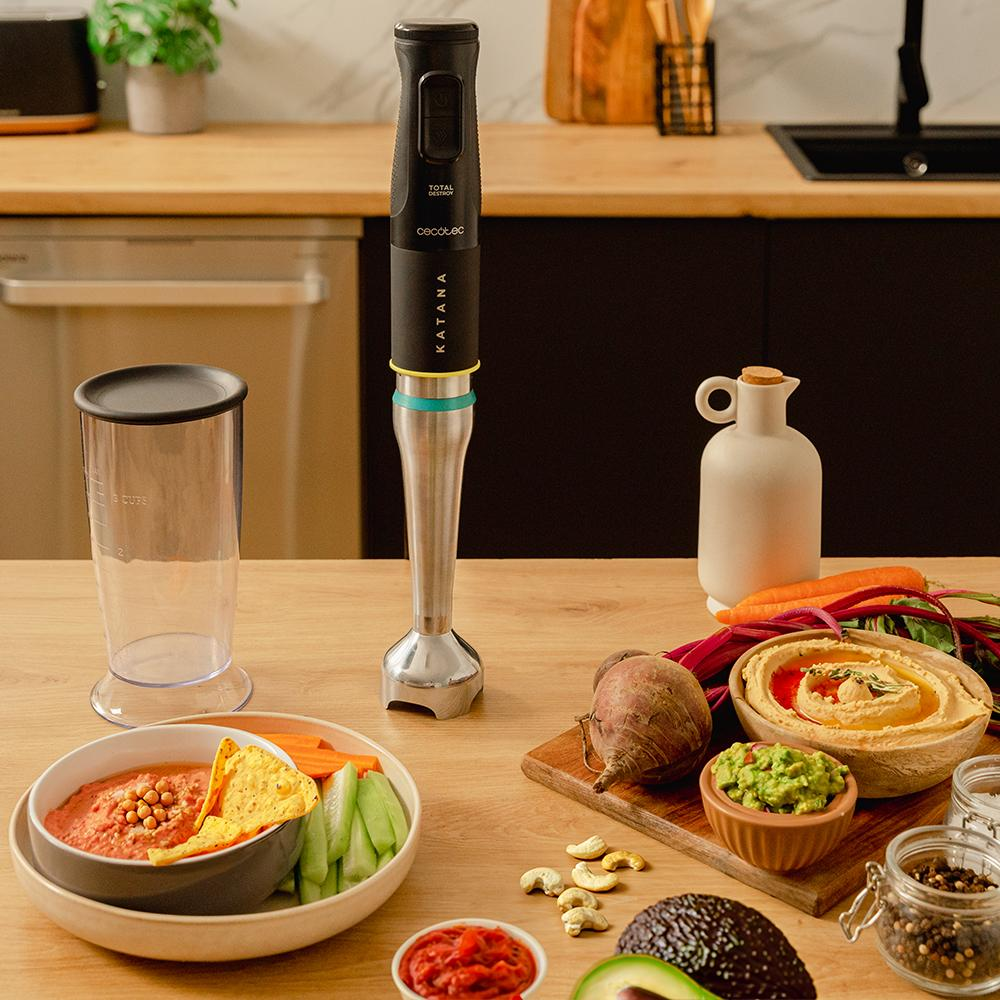 Una batidora está sobre una mesa de madera con hummus, verduras y salsa. A la izquierda, una taza medidora transparente.