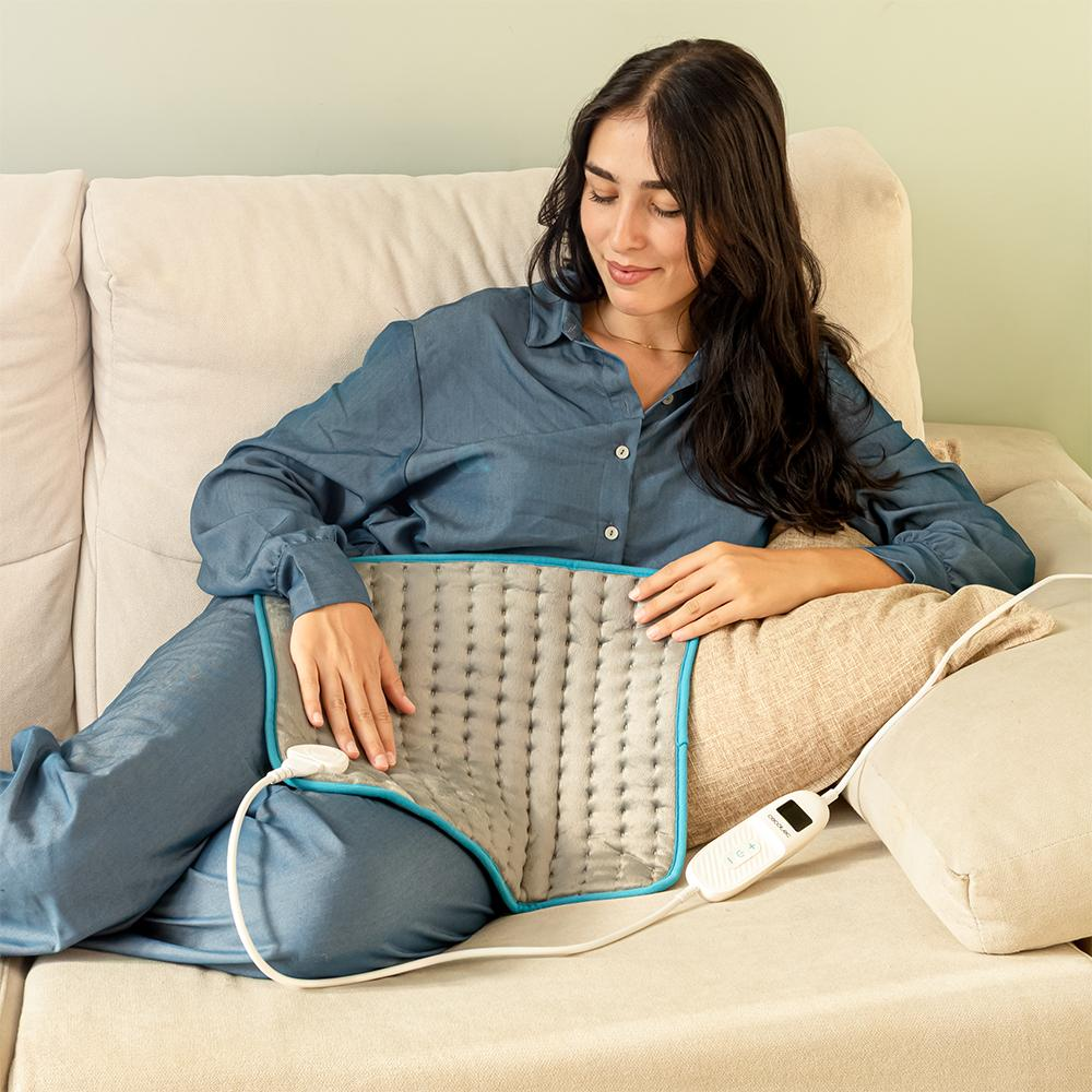 Mujer con ropa azul en un sofá usando una almohadilla térmica. Ella está sonriendo. Panel de control blanco visible. Cojines y sofá beige.