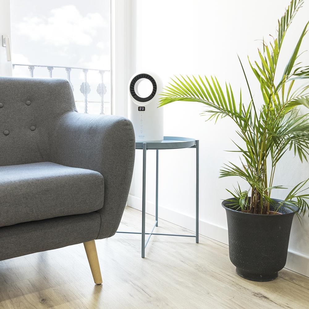 Un sillón gris, un ventilador blanco sobre una mesita, una planta en maceta y una ventana con balcón están en la habitación.