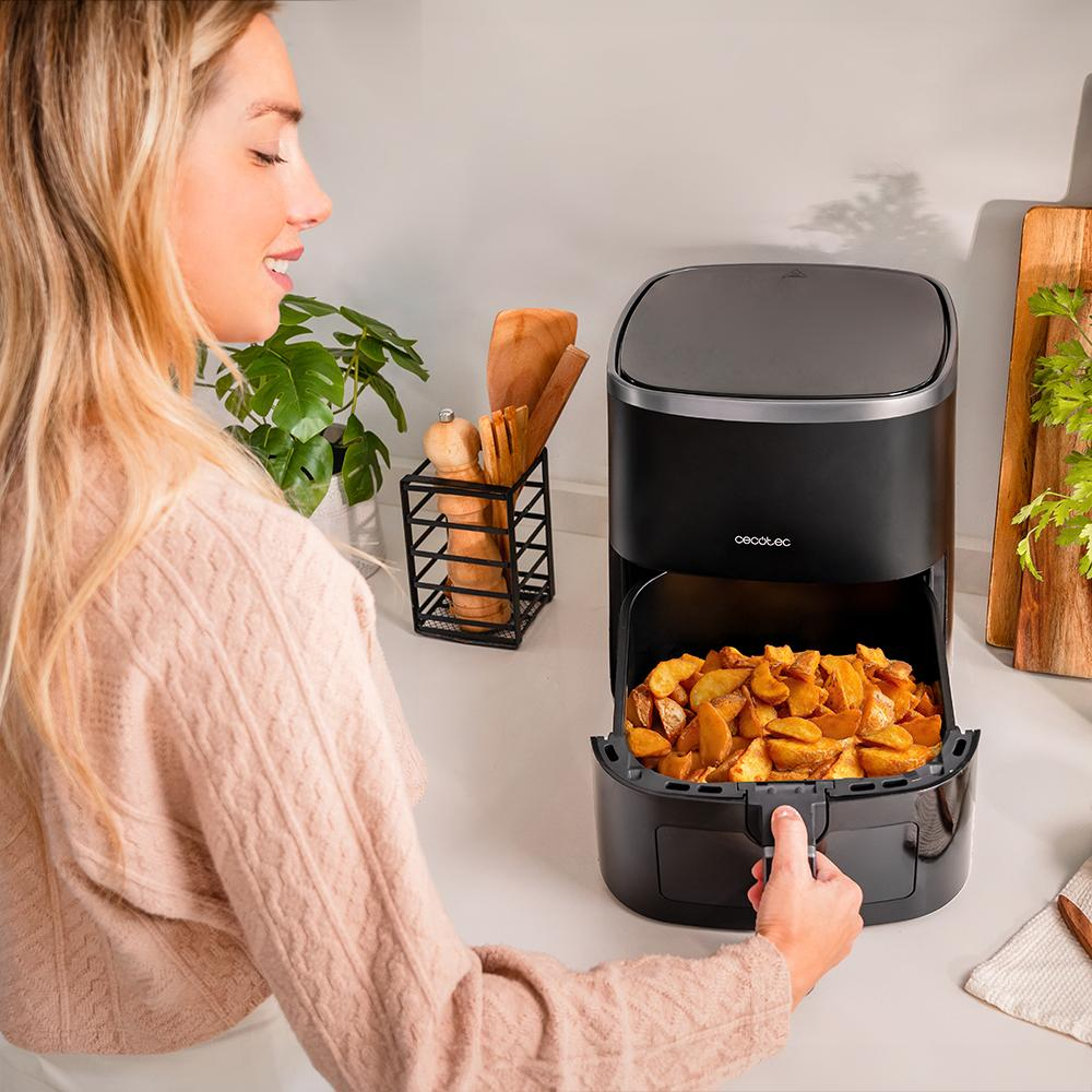 Mujer rubia opera una freidora de aire con gajos de patata. Ambiente de cocina con utensilios de madera y vegetación.