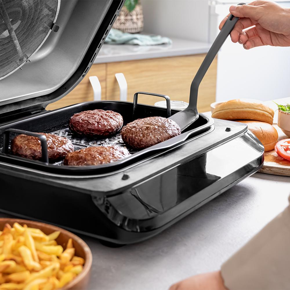 Hamburguesas a la parrilla en un electrodoméstico negro. Una mano sostiene una espátula. Papas fritas y pan de hamburguesa en el fondo.