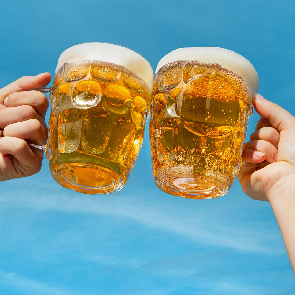 Dos manos sostienen jarras de cerveza chocando contra un cielo azul. Las jarras están llenas de cerveza y cubiertas de espuma.