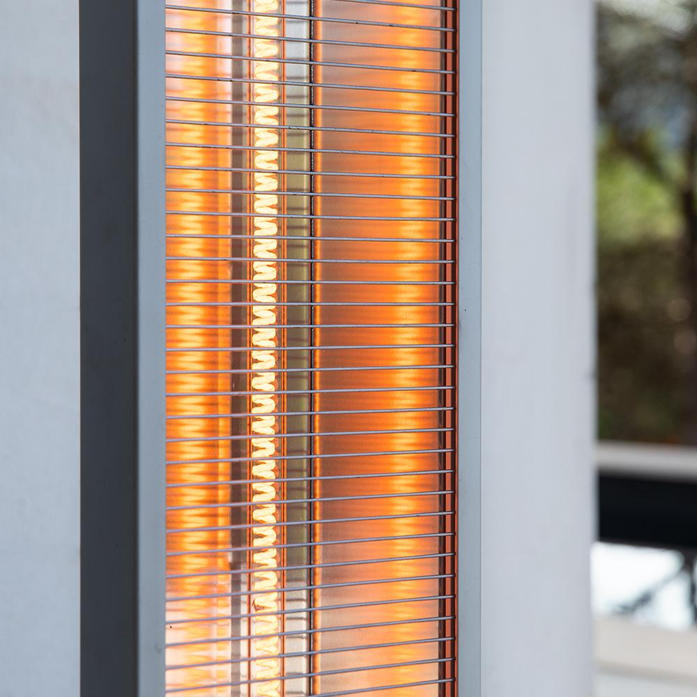 Primer plano de un calefactor de patio. Tiene una rejilla de metal, un brillo anaranjado en el interior y un marco blanco. El fondo tiene una pared blanca.