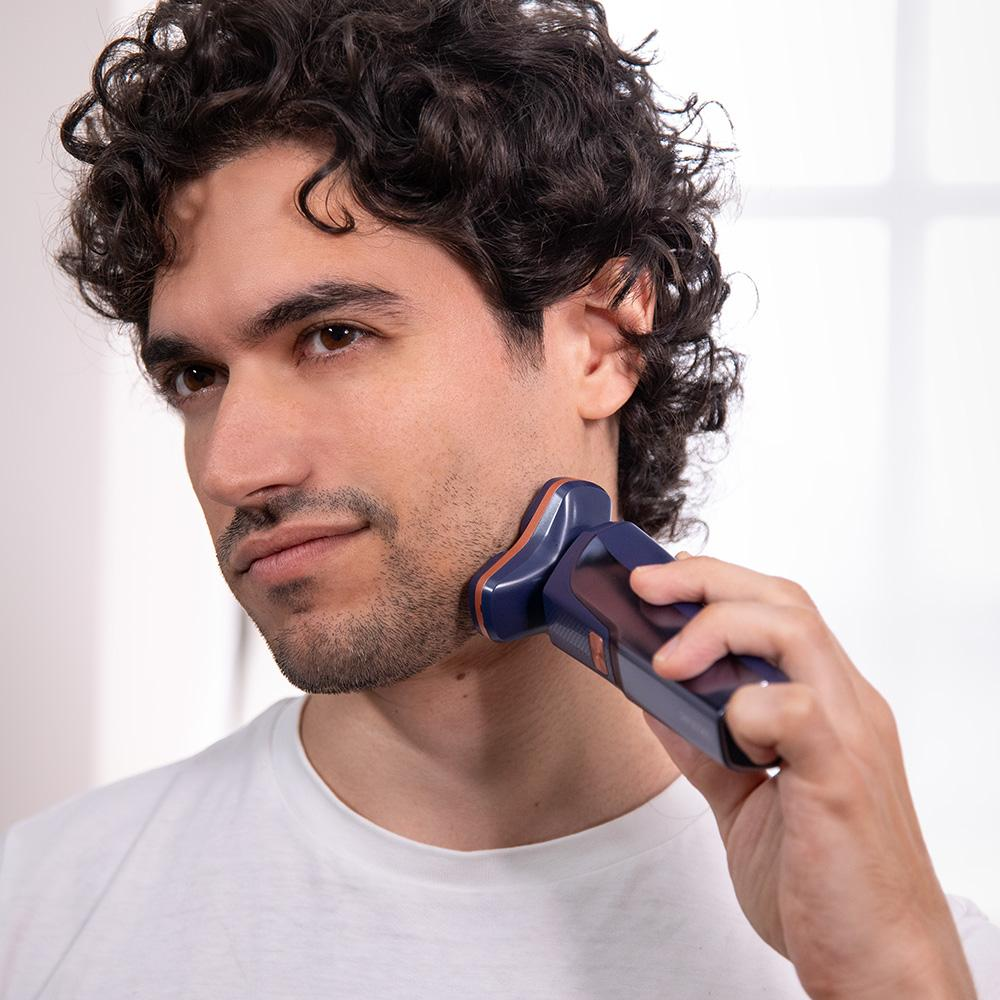 Un hombre se afeita la barba con una afeitadora eléctrica. Lleva una camiseta blanca y tiene el pelo rizado oscuro. La afeitadora es azul marino y cobriza.