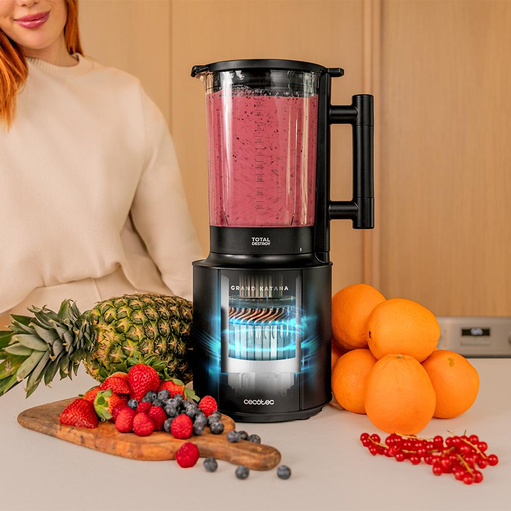 Una licuadora con líquido rosa está junto a fruta: piña, fresas, naranjas y bayas. Una mujer está cerca.