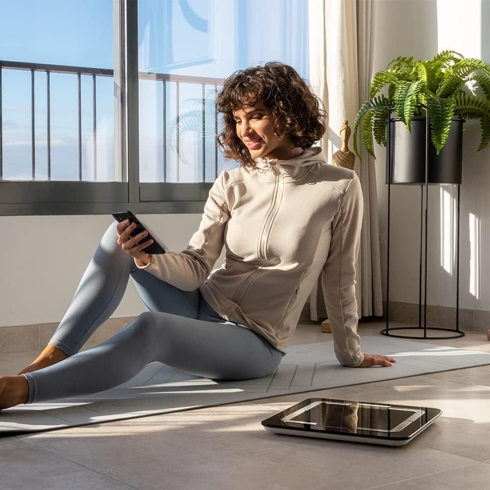 Una mujer sentada en una esterilla mira su teléfono. Hay una báscula frente a ella. Un helecho en el fondo.