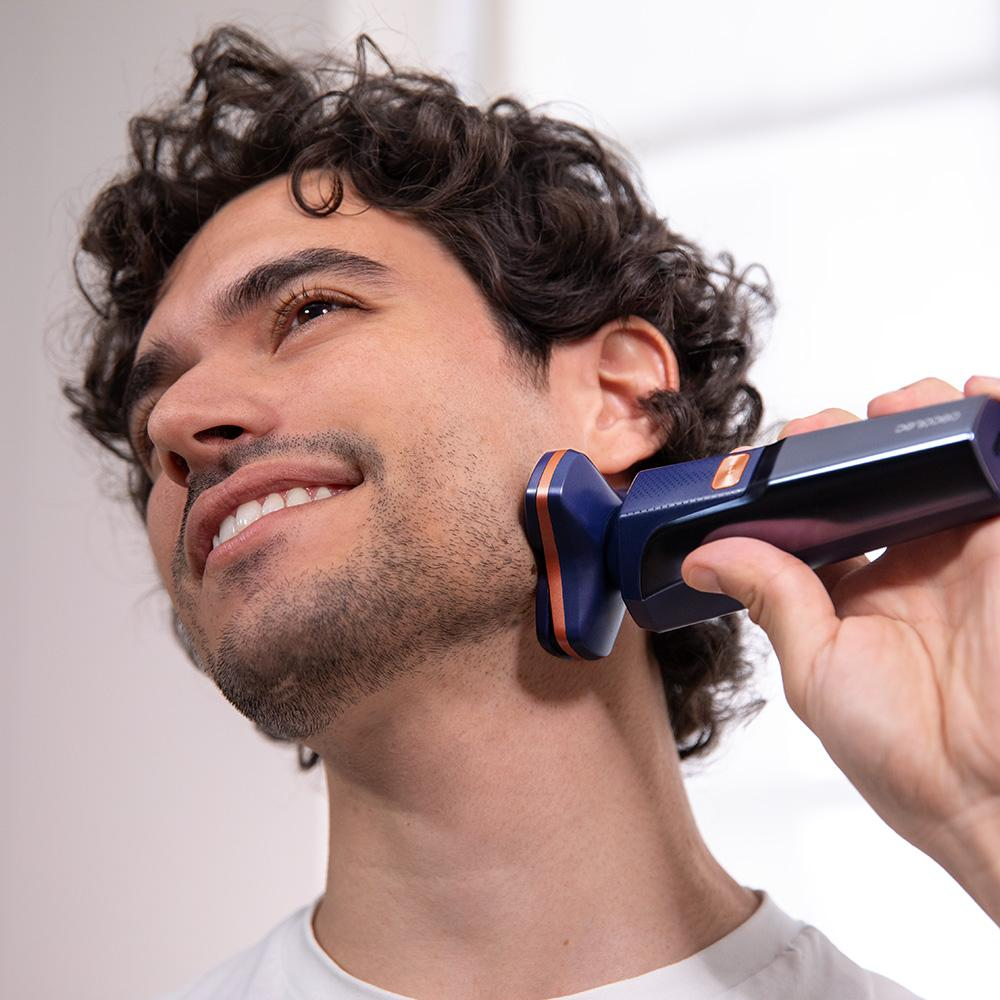 Un hombre se afeita la barba con una afeitadora eléctrica. La afeitadora es azul y naranja. El hombre está sonriendo.