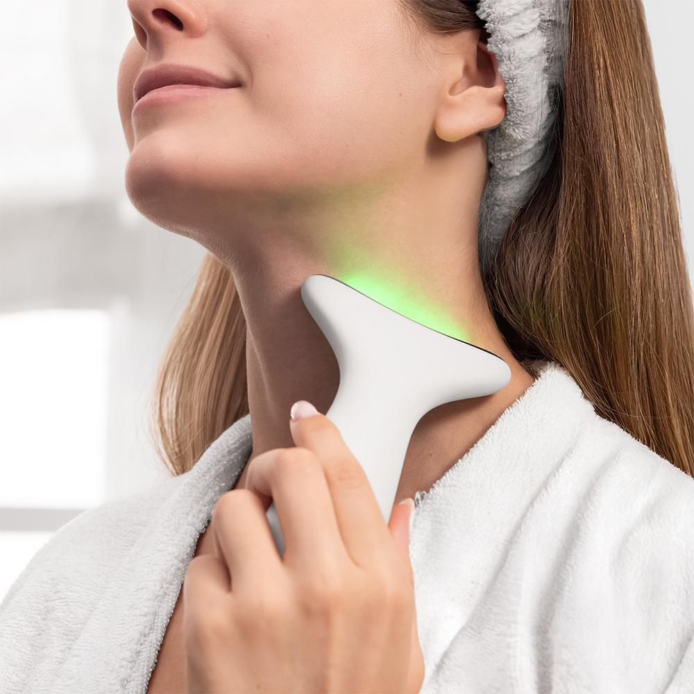 Una mujer aplica un dispositivo de belleza blanco en su cuello. El dispositivo emite una luz verde. Lleva una bata blanca y una diadema.