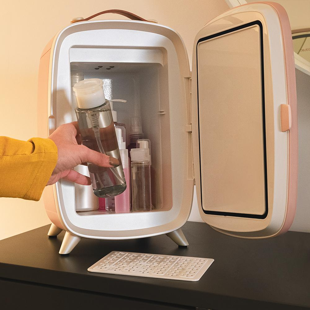 Una mano con manga amarilla sostiene una botella transparente dentro de una nevera rosa. Se ven otras botellas. La puerta de la nevera está abierta.