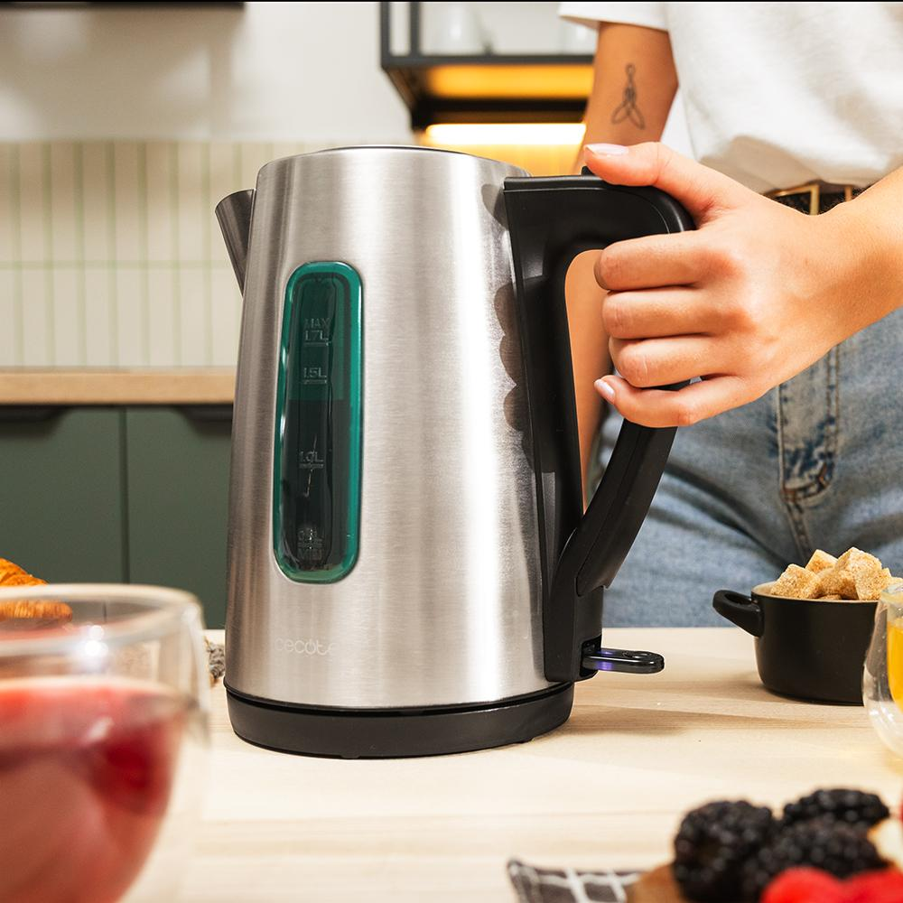 Una persona sostiene una tetera eléctrica de acero inoxidable. Un vaso de jugo y moras en primer plano.