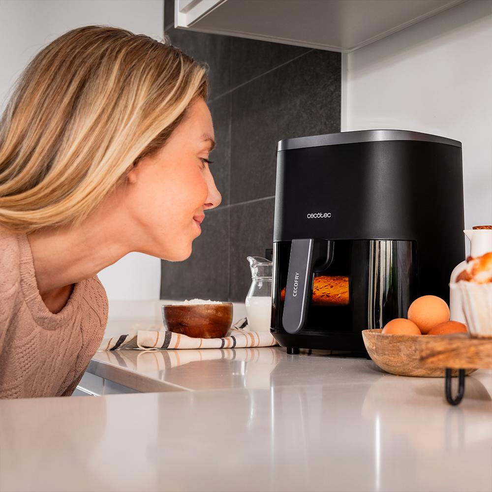 Mujer mirando una freidora de aire negra. Huevos, un cuenco de harina y leche están en la encimera. El fondo es una cocina.