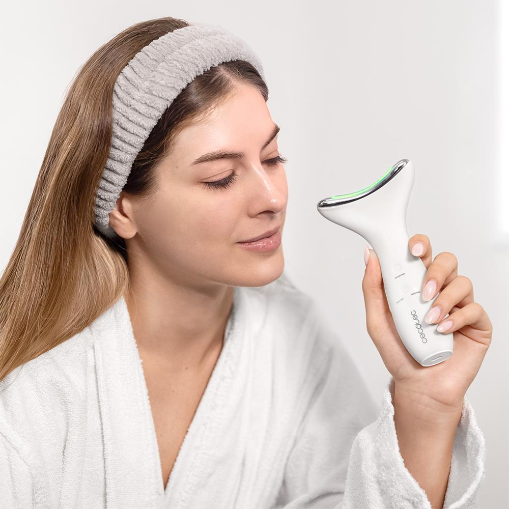 Mujer en bata sostiene un dispositivo de belleza blanco. Lleva una diadema y mira hacia abajo. El dispositivo tiene luces verdes.