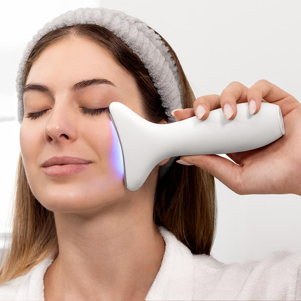 Mujer usa un dispositivo facial blanco debajo del ojo. Lleva una diadema y bata. El dispositivo emite una luz azul.