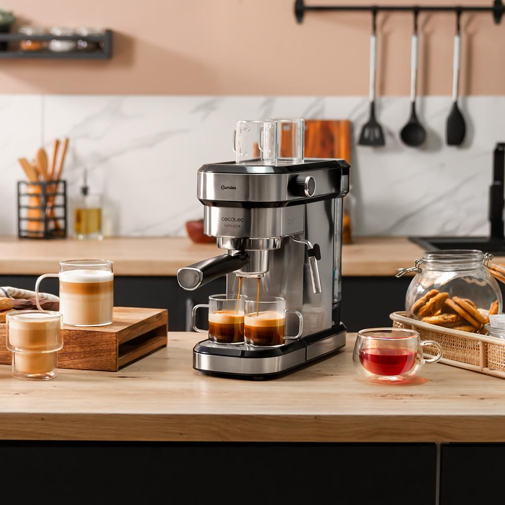 Máquina de espresso vertiendo café en dos tazas de vidrio. Se ven otras tazas con café y té, galletas y utensilios de cocina.