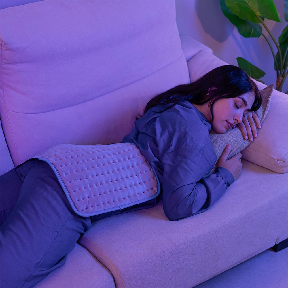 Una mujer descansa en un sofá con una almohadilla térmica. Está dormida, con la cabeza sobre una almohada. La escena está iluminada en morado.