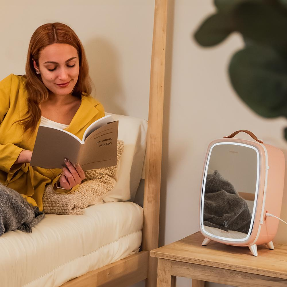 Mujer en chaqueta amarilla lee libro en cama. Mini-nevera rosa con espejo sobre mesa de madera.