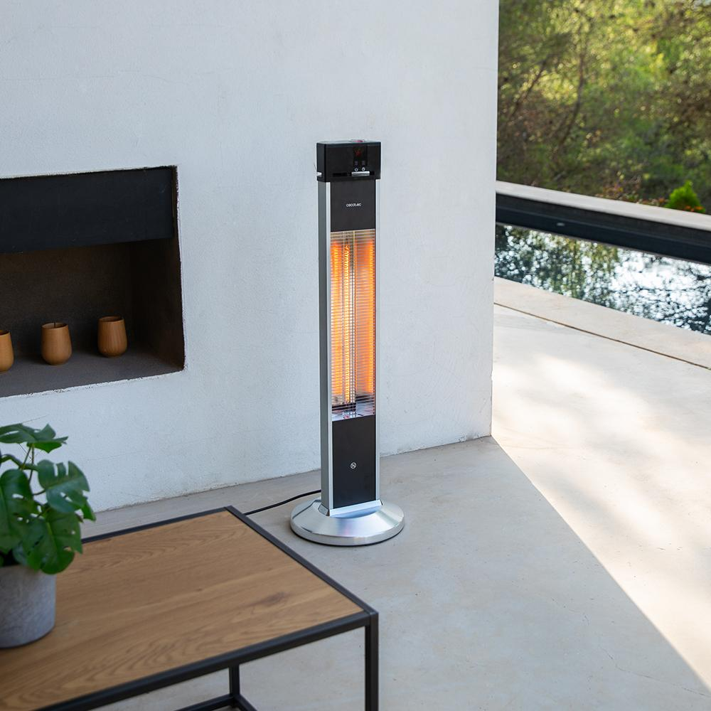 Un calefactor exterior está en un patio. Tiene una parte superior negra, cuerpo plateado y un resplandor naranja. Una mesa y plantas están en primer plano.