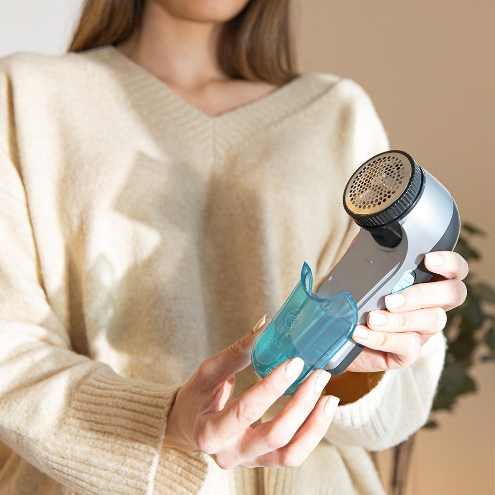 Una mujer sostiene una afeitadora de tela. La afeitadora es gris y negra, con un colector de pelusas azul. Lleva un suéter beige.