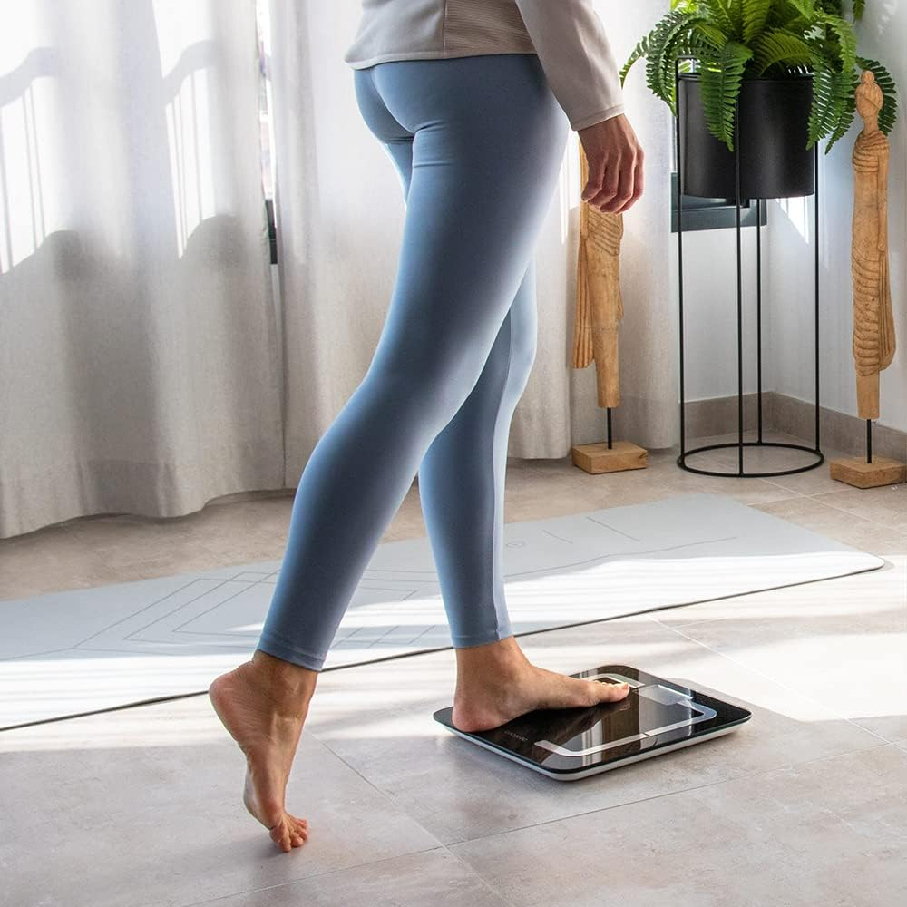 Una persona está de pie sobre una báscula en una habitación. Lleva mallas azules y una camisa clara. Una esterilla de yoga y plantas están al fondo.