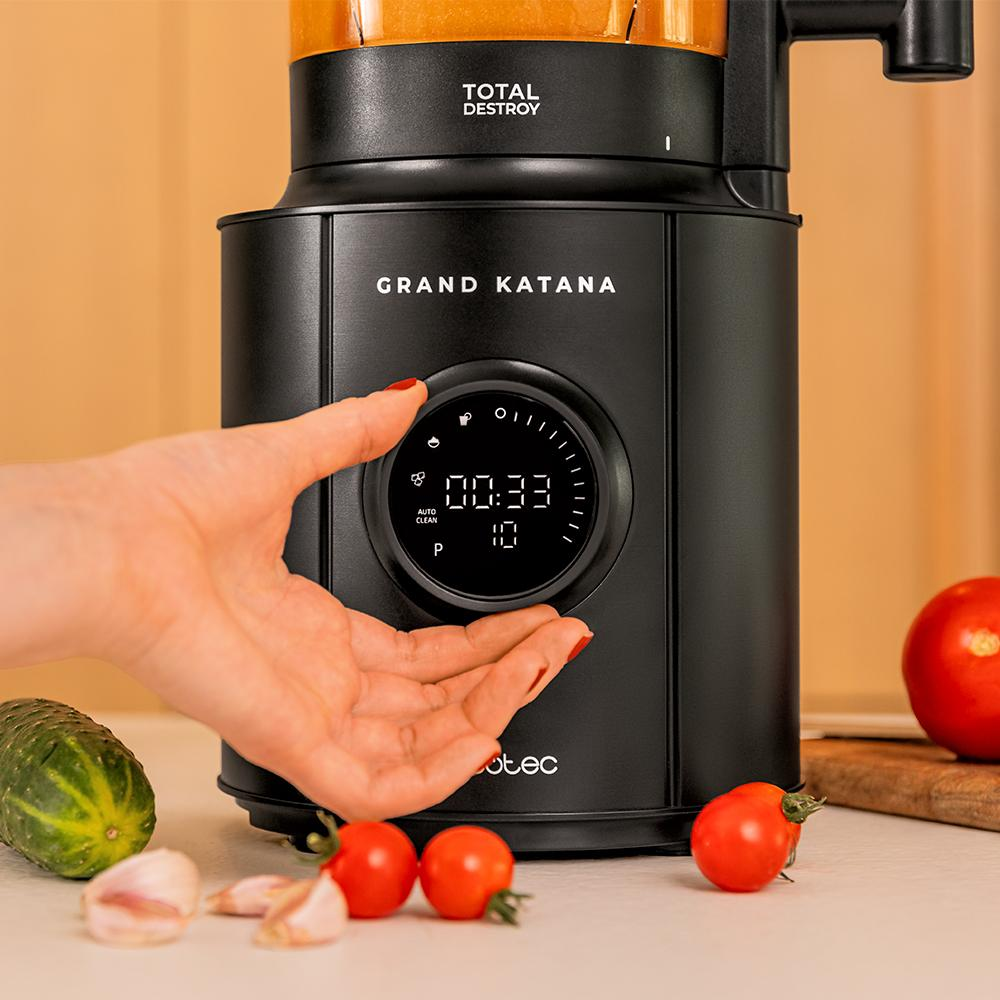 Una batidora negra con las palabras GRAND KATANA. Una mano está a punto de tocar la pantalla de la batidora. Tomates y ajo sobre la mesa.
