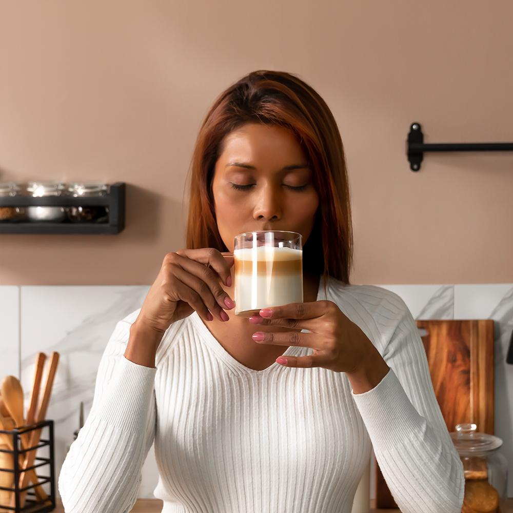 Mujer de blanco bebe café en capas, ojos cerrados. Ambiente de cocina con madera, especiero y mármol. Pared marrón claro.
