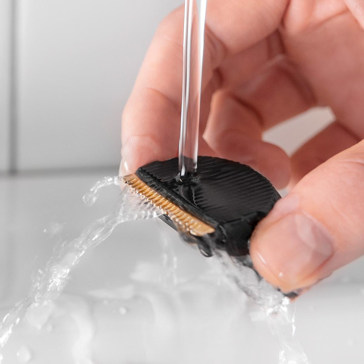 Una mano sostiene un cabezal de afeitadora negro que se enjuaga con agua. La cuchilla es dorada.