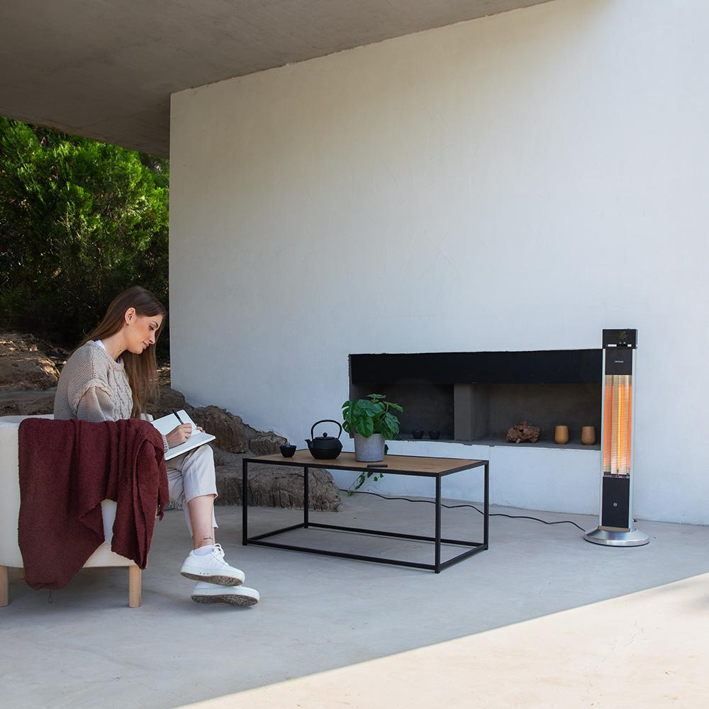 Mujer escribiendo en un patio. Hay una mesa con tetera, planta y calefactor. Una chimenea y una pared blanca al fondo.
