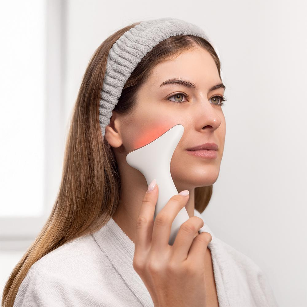 Una mujer usa un dispositivo facial blanco en su mejilla. Lleva una diadema gris y una bata blanca. Su mejilla tiene un brillo rojo.